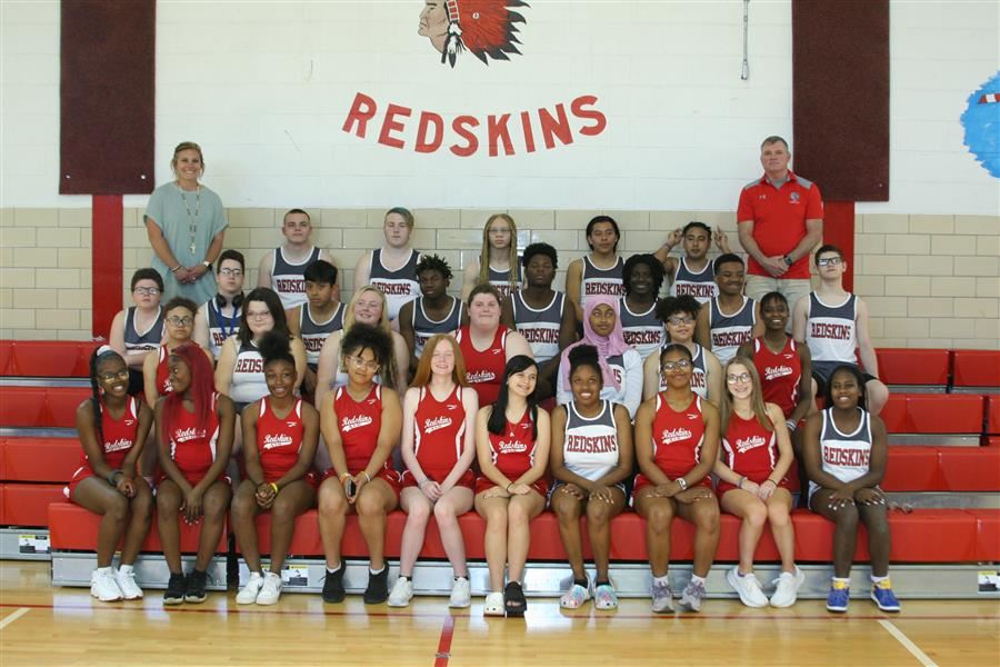  Members of the boys & girls track teams smile to camera while seated in gym bleachers with coaches.